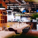 Stylish restaurant interior with an open kitchen where a chef works, laid tables and colorful designer chairs.