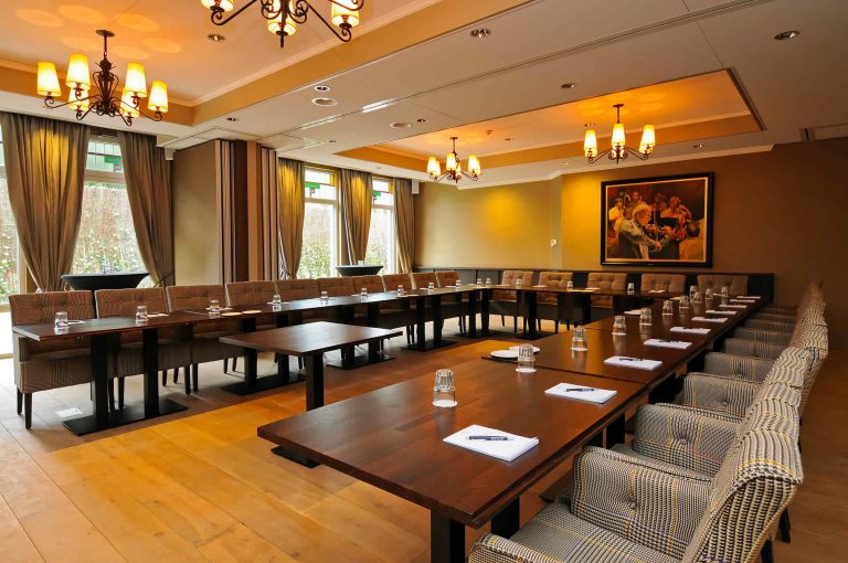 U-vormige houten vergadertafel met glazen en notitieblokjes in een sfeervolle conferentiezaal met patroonstoelen en kroonluchters.