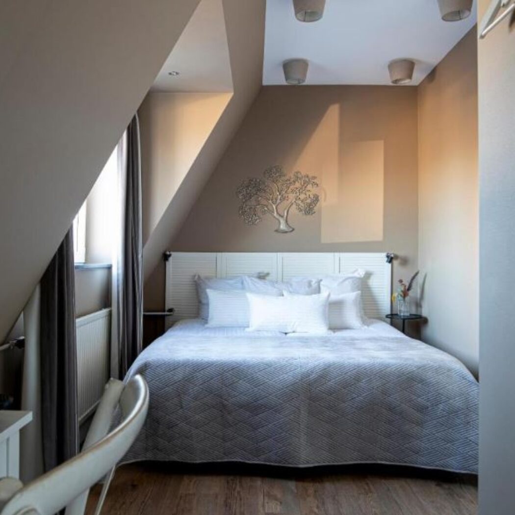 Knusse zolderkamer met wit bed, grijs dekbed, houten vloer en een decoratieve zilveren boom aan de muur.