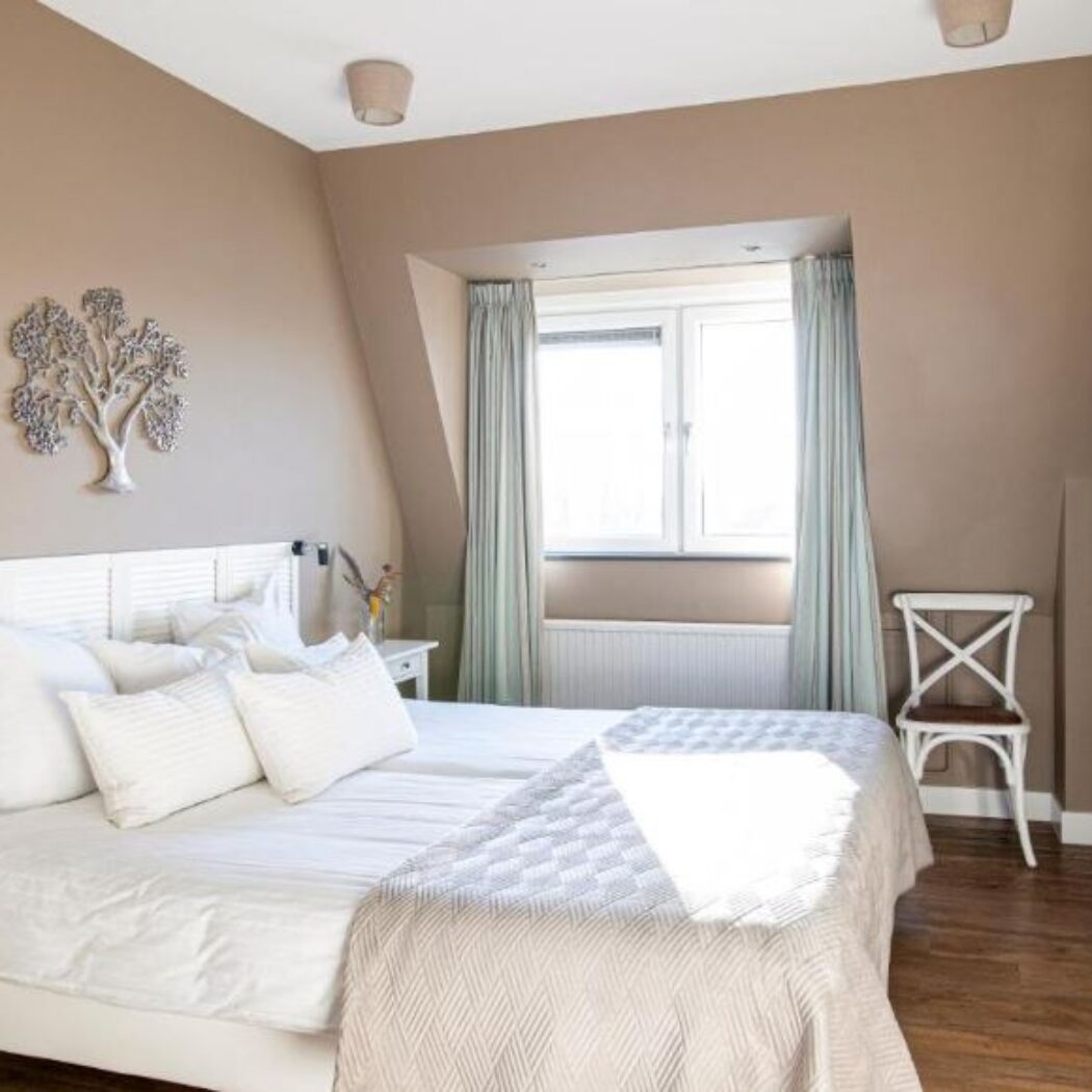 Lichte slaapkamer met wit tweepersoonsbed, zandkleurige muren, raam, witte stoel en zilveren boomdecoratie.