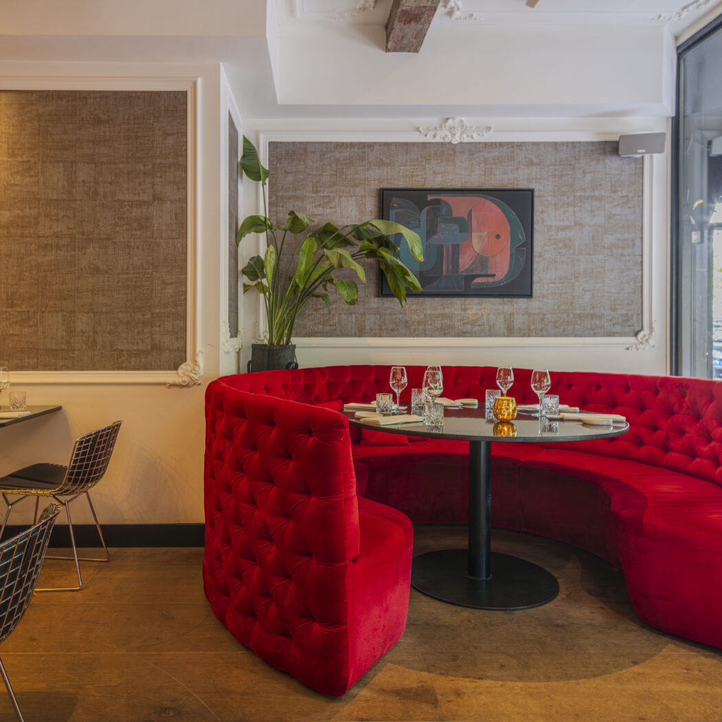 Stijlvol restaurantinterieur met een grote, gebogen rode fluwelen zitbank, gedekte tafels en houten vloer.