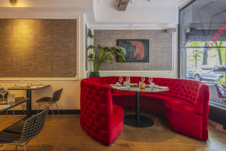 Stijlvol restaurantinterieur met een grote, gebogen rode fluwelen zitbank, gedekte tafels en houten vloer.