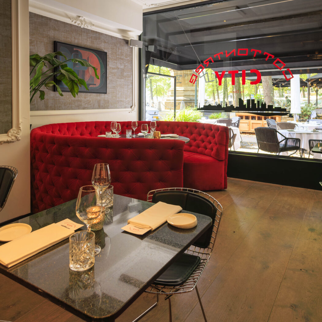Modern restaurant interieur met rood bankstel, zwarte tafels en uitzicht op het terras; CITY op het raam.