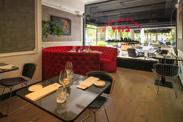 Modern restaurant interieur met rood bankstel, zwarte tafels en uitzicht op het terras; CITY op het raam.