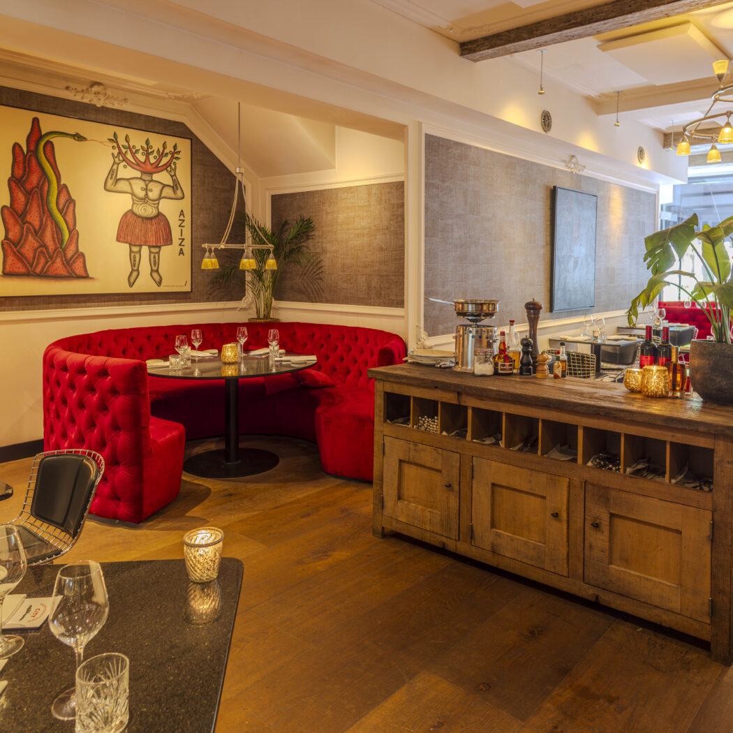 Gezellig restaurant interieur met rode ronde zithoek, houten vloer, kunstwerk AZIZA en een houten counter met culinaire items.