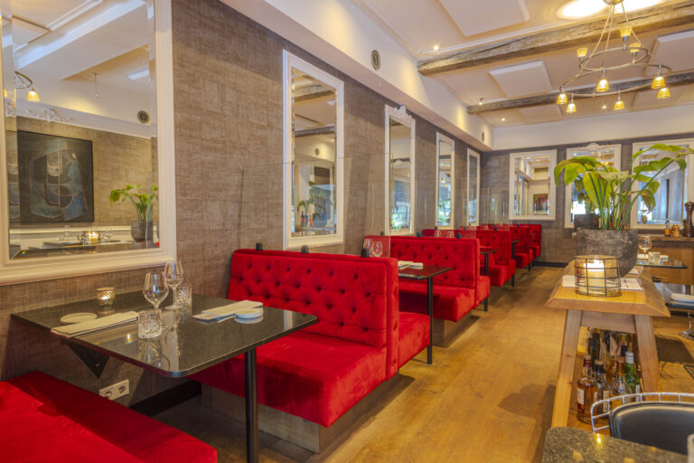 Interieur van een restaurant met rode fluwelen banken, zwarte tafels, wandspiegels en houten vloer.