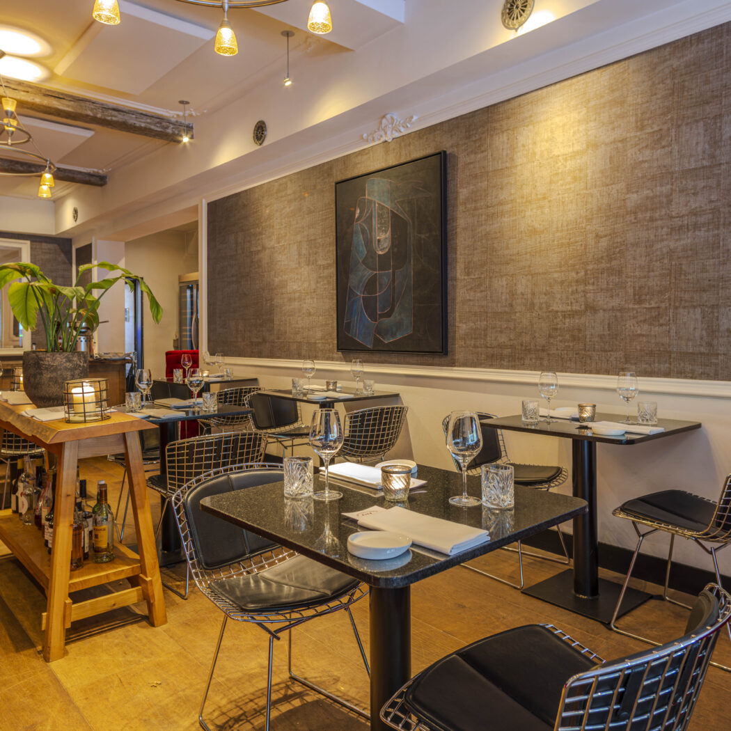 Sfeervol restaurantinterieur met zwarte granieten tafels, draadstoelen, houten vloer, planten en abstracte kunst.