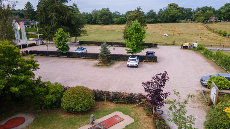 Landelijke gravel parkeerplaats met autos, hagen, bomen en een minigolfbaan, met een paard in de weide.