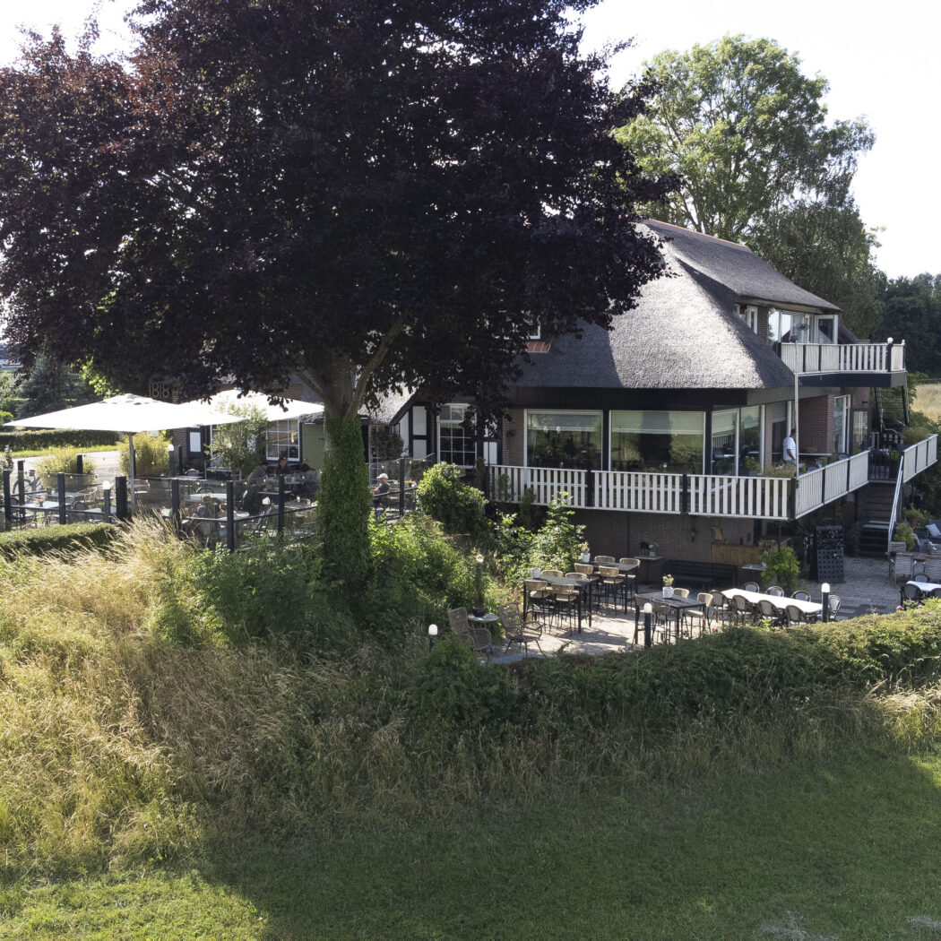 Rietgedekt restaurant met buitenterrassen en witte balkons, deels onder een grote roodbladige boom.