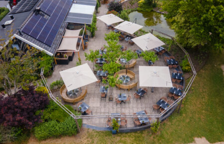 Luchtfoto van restaurantterras met beige parasols, tafels en stoelen, een gebouw met zonnepanelen en vijver.