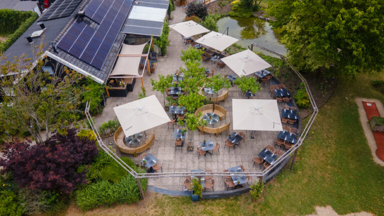 Luchtfoto van restaurantterras met beige parasols, tafels en stoelen, een gebouw met zonnepanelen en vijver.