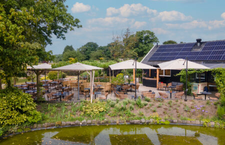 Ruim buitenterras met rotan stoelen, tafels en beige parasols aan een vijver, naast een gebouw met zonnepanelen.