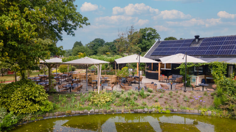 Ruim buitenterras met rotan stoelen, tafels en beige parasols aan een vijver, naast een gebouw met zonnepanelen.