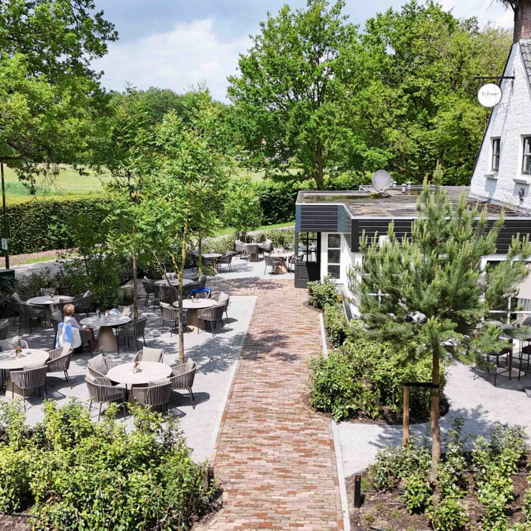 Uitgestrekt restaurantterras met ronde tafels, omringd door groen en een bakstenen pad, bij gebouw De Hoppe.