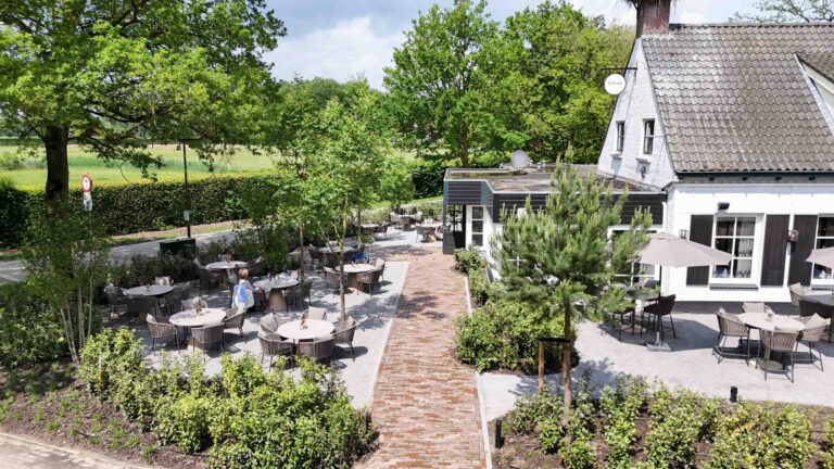 Uitgestrekt restaurantterras met ronde tafels, omringd door groen en een bakstenen pad, bij gebouw De Hoppe.