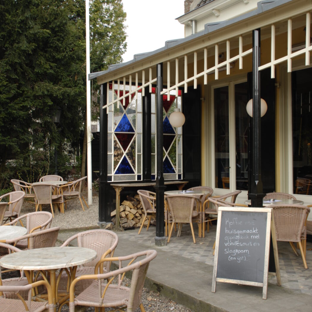 Terras van een café met rieten stoelen en tafels, krijtbord met huisgemaakt appelgebak en glas-in-loodramen.
