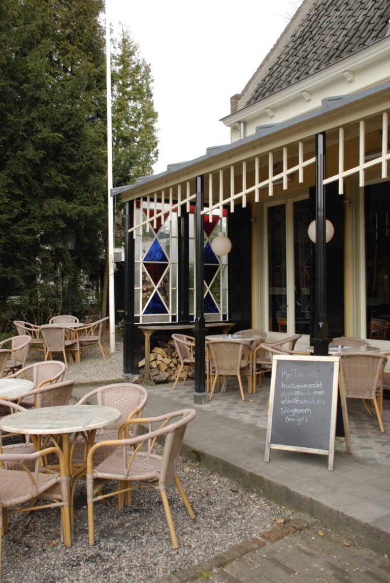 Terras van een café met rieten stoelen en tafels, krijtbord met huisgemaakt appelgebak en glas-in-loodramen.