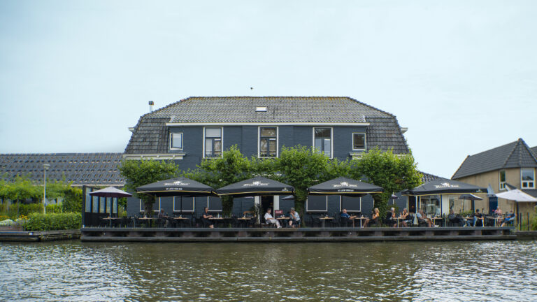 Donkerblauw gebouw met grijs dak en terras aan het water, met mensen onder zwarte Hertog Jan parasols.
