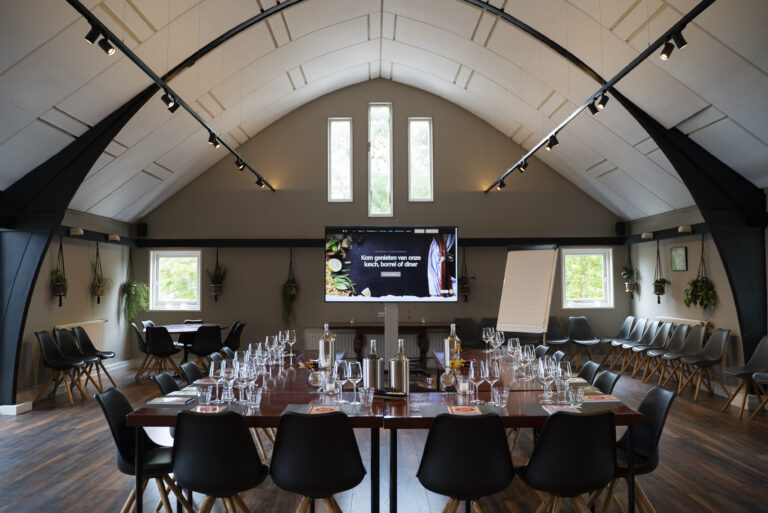 Ruime evenementenlocatie met hoog boogplafond, lange houten tafel, glazen en scherm: Kom genieten van onze lunch, borrel of diner.