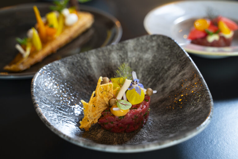 Fijnproevers steak tartaar met eidooier, blauwe bloem en krokante crunch op een donkergrijs bord.