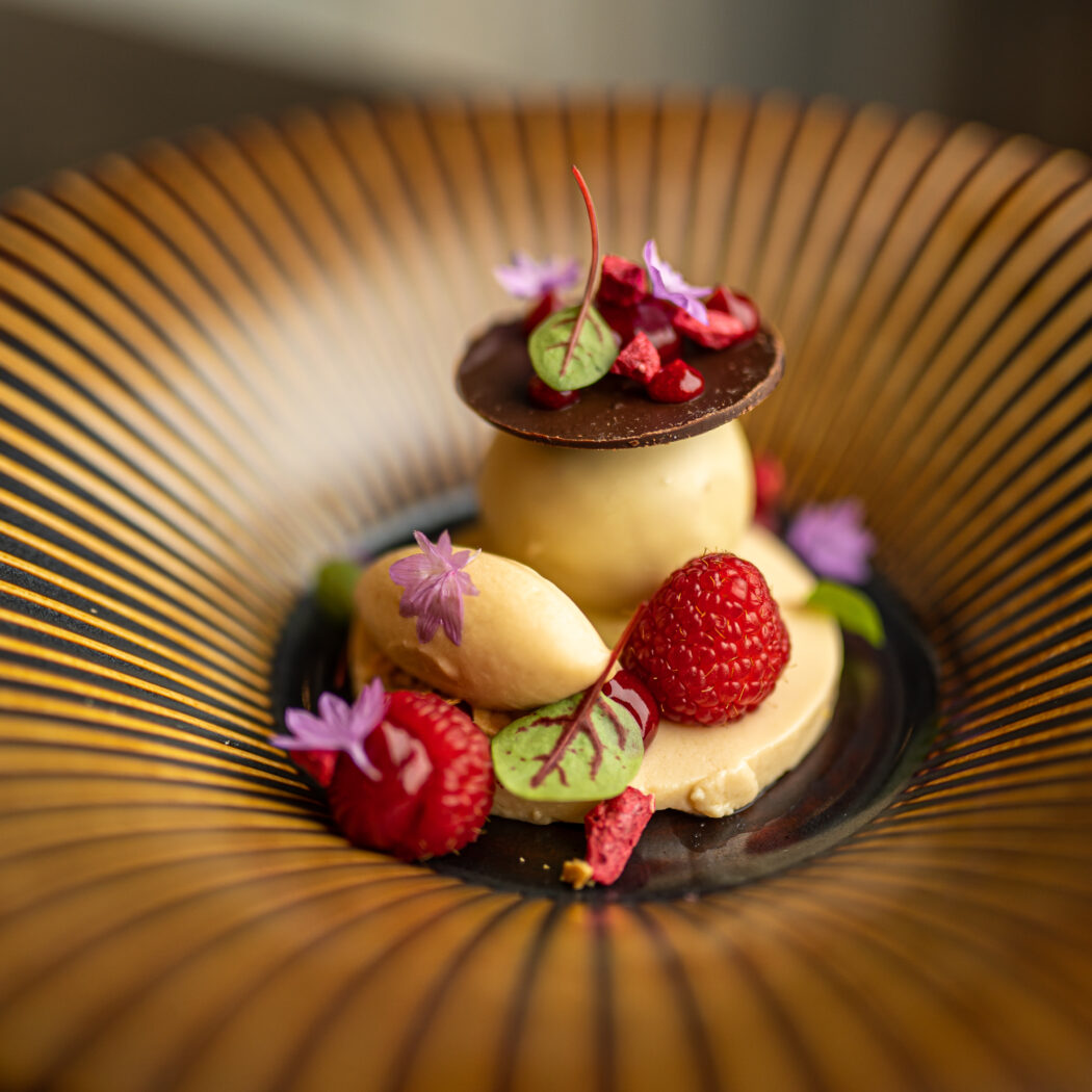 Elegant nagerecht met crème dessert, frambozen, chocolade schijf, en paarse bloemetjes op een goudkleurig geribbeld bord.
