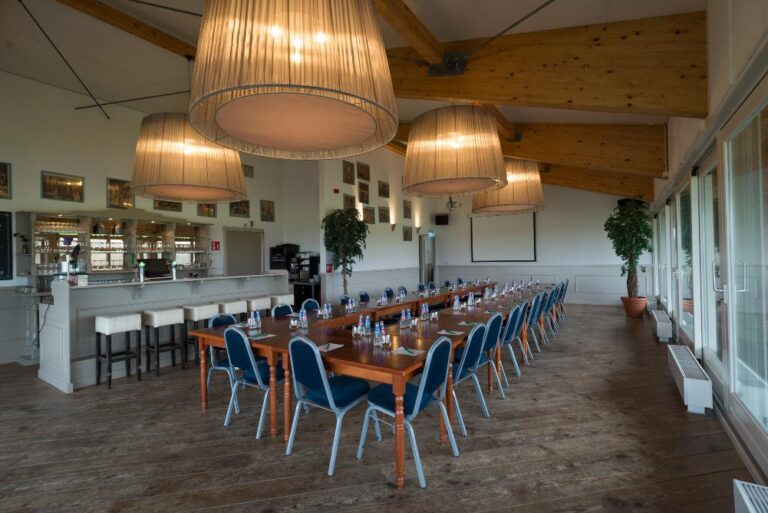 Ruime vergaderlocatie met lange houten tafel, blauwe stoelen, bar, projectiescherm en grote hanglampen.