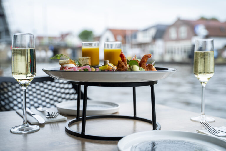 Een feestelijk plateau met hapjes, twee glazen champagne en jus dorange op een buitentafel aan het water.