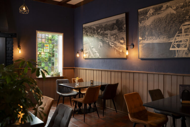 Restaurantinterieur met donkerblauwe muren, houten lambrisering, raam met glas-in-lood, tafels, stoelen en historische zwart-wit fotos.