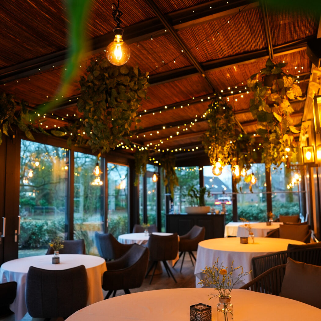 Gezellige serre van een restaurant met bamboe dak, ronde tafels, hangplanten en warme decoratieve verlichting.