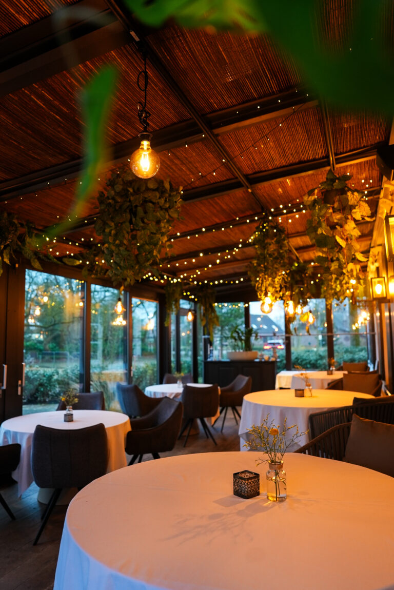 Gezellige serre van een restaurant met bamboe dak, ronde tafels, hangplanten en warme decoratieve verlichting.