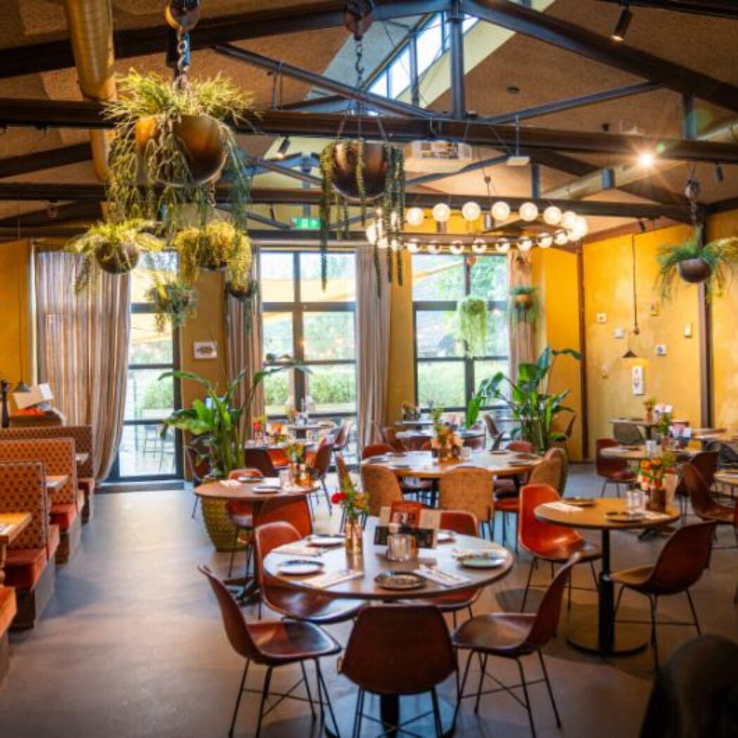 Gezellig restaurantinterieur met gele muren, veel planten, ronde tafels en oranje banken bij grote ramen.