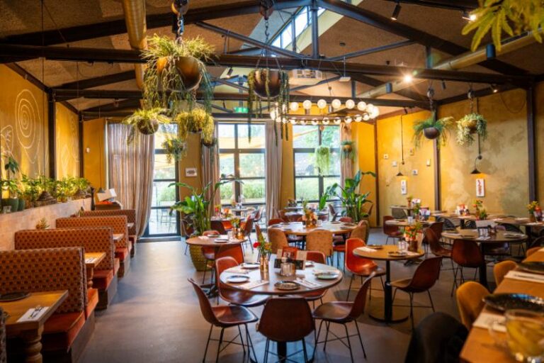 Gezellig restaurantinterieur met gele muren, veel planten, ronde tafels en oranje banken bij grote ramen.