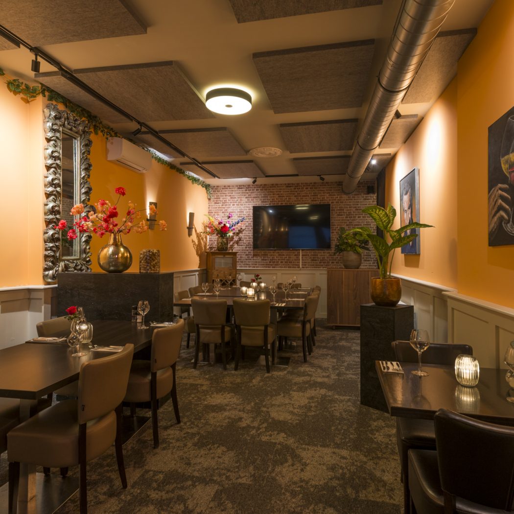 Sfeervol restaurantinterieur met warm oranje muren, gedekte eettafels, bloemstukken en kunst aan de muur.