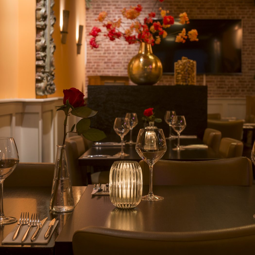 Restauranttafel gedekt met gloeiend kaarslicht, rode roos en wijnglazen, in een warme, intieme sfeer.