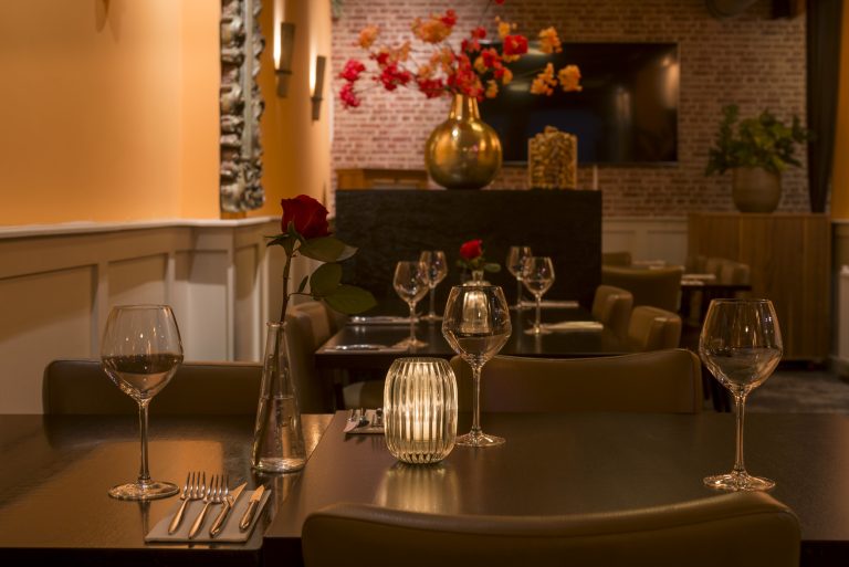 Restauranttafel gedekt met gloeiend kaarslicht, rode roos en wijnglazen, in een warme, intieme sfeer.