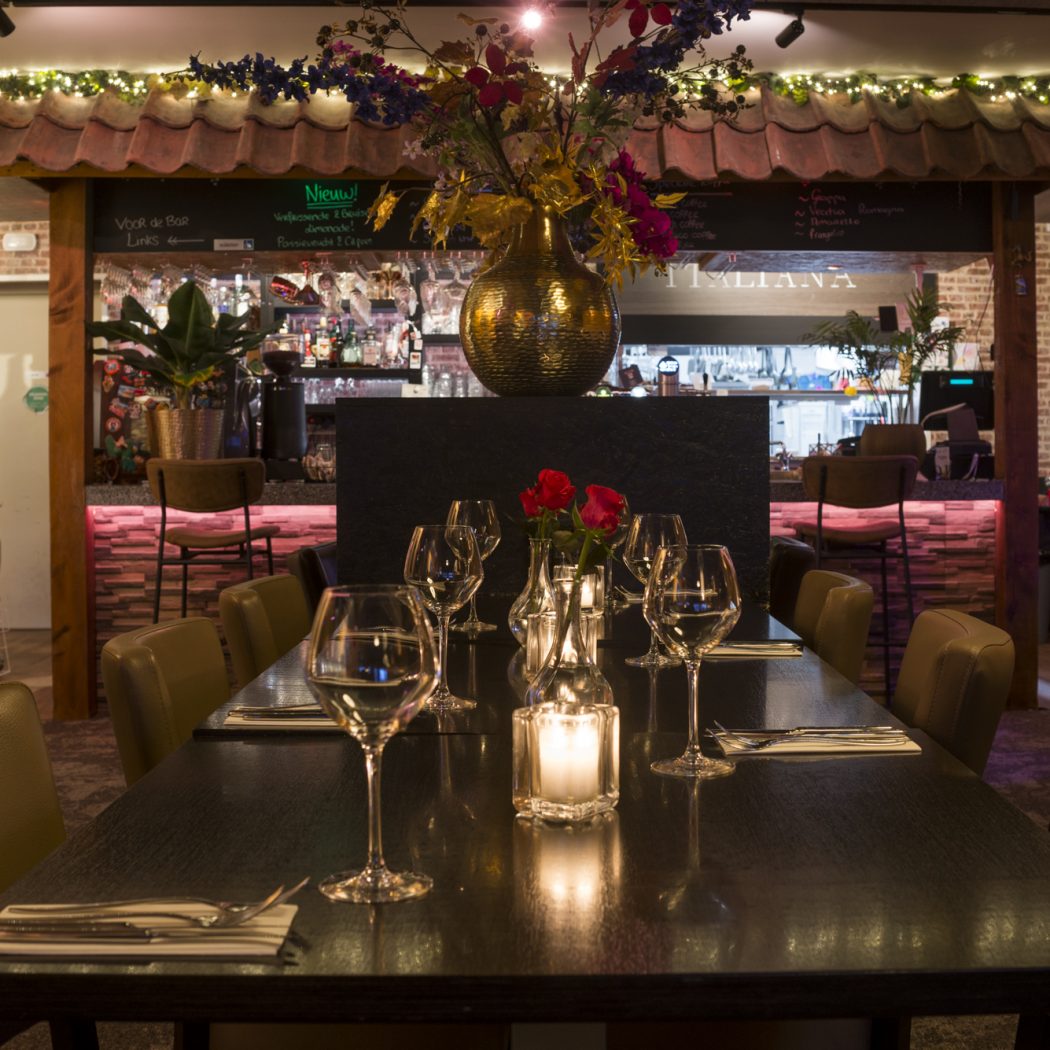 Lange eettafel in een sfeervol Italiaans restaurant, gedekt met glazen en kaarslicht, met een verlichte bar op de achtergrond.