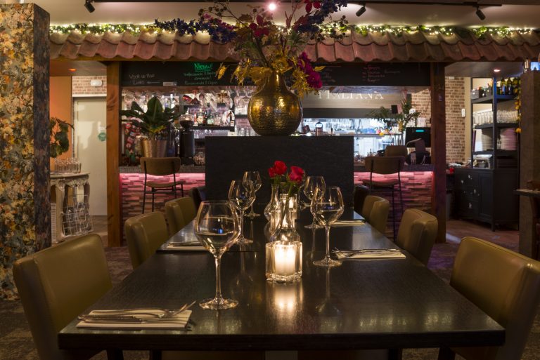Lange eettafel in een sfeervol Italiaans restaurant, gedekt met glazen en kaarslicht, met een verlichte bar op de achtergrond.
