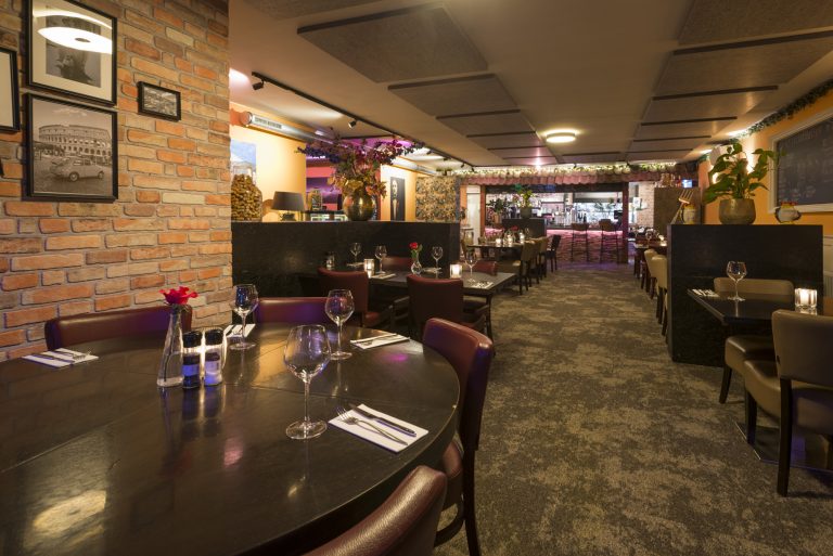 Sfeervol restaurant met gedekte tafels, bakstenen muur met Rome-afbeeldingen en een verlichte bar achterin.