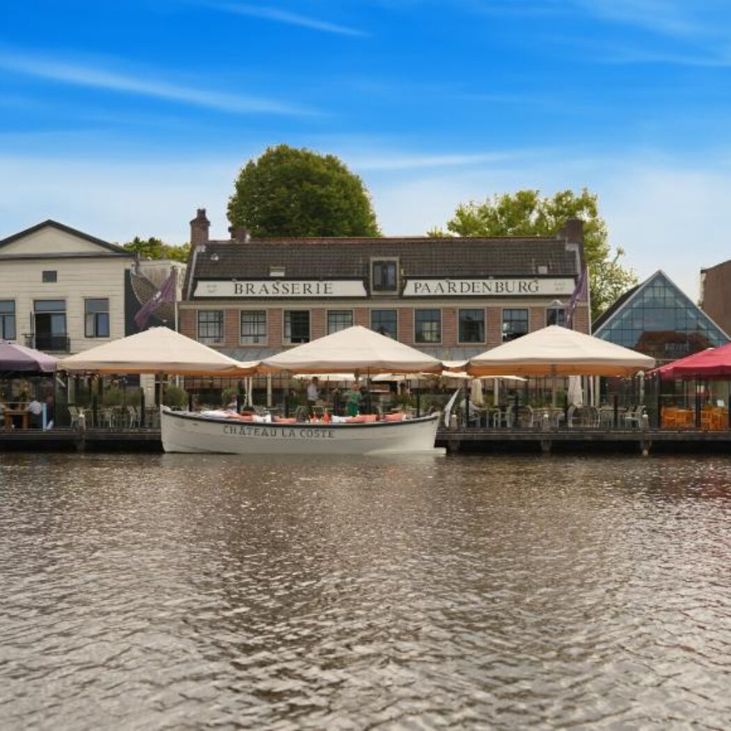 Uitzicht op een kanaal met terrassen vol mensen onder kleurrijke parasols, een witte boot Chateau La Coste en de gevel van Brasserie Paardenburg.