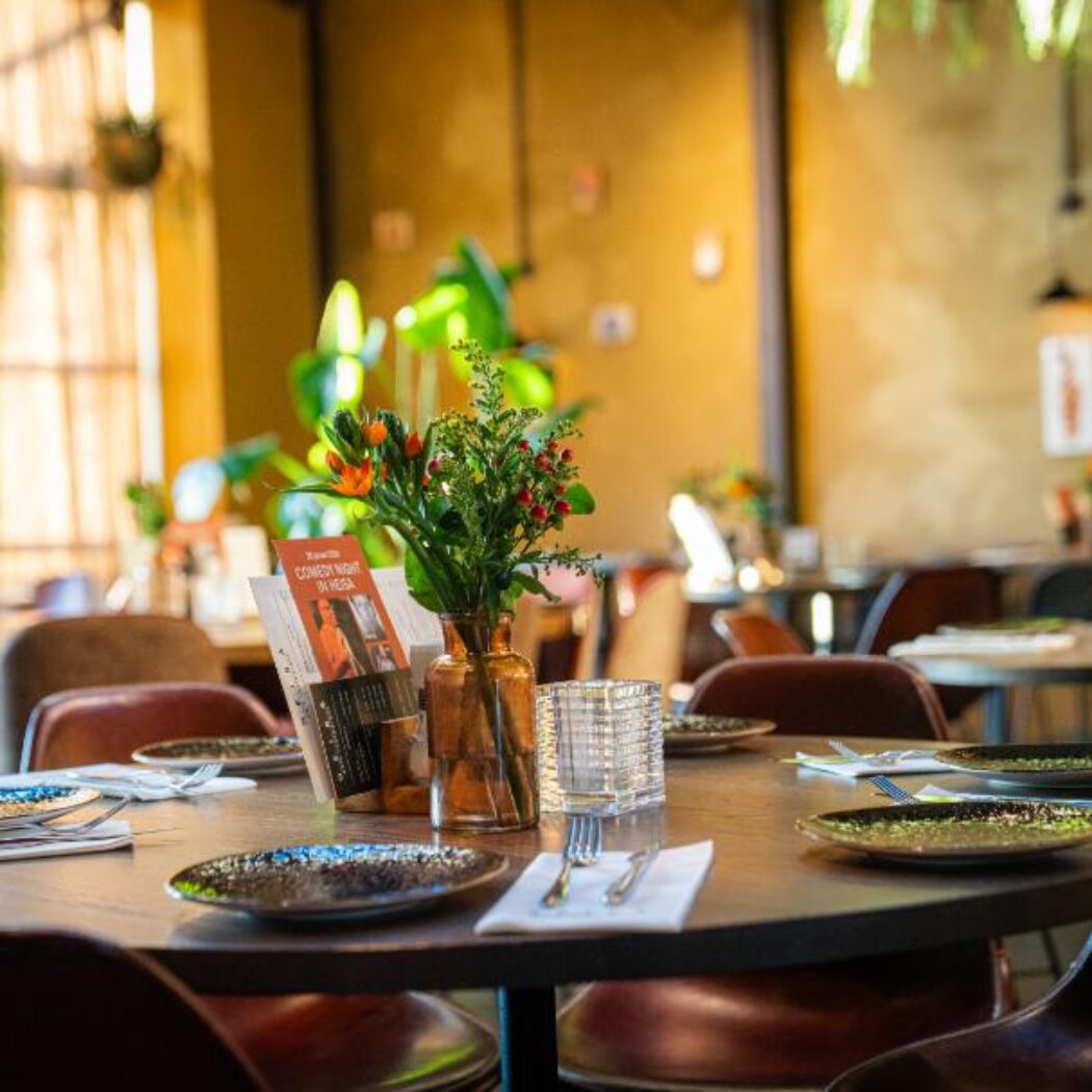 Sfeervolle restauranttafel gedekt met donkere borden, bestek, oranje bloemen en een COMEDY NIGHT flyer.