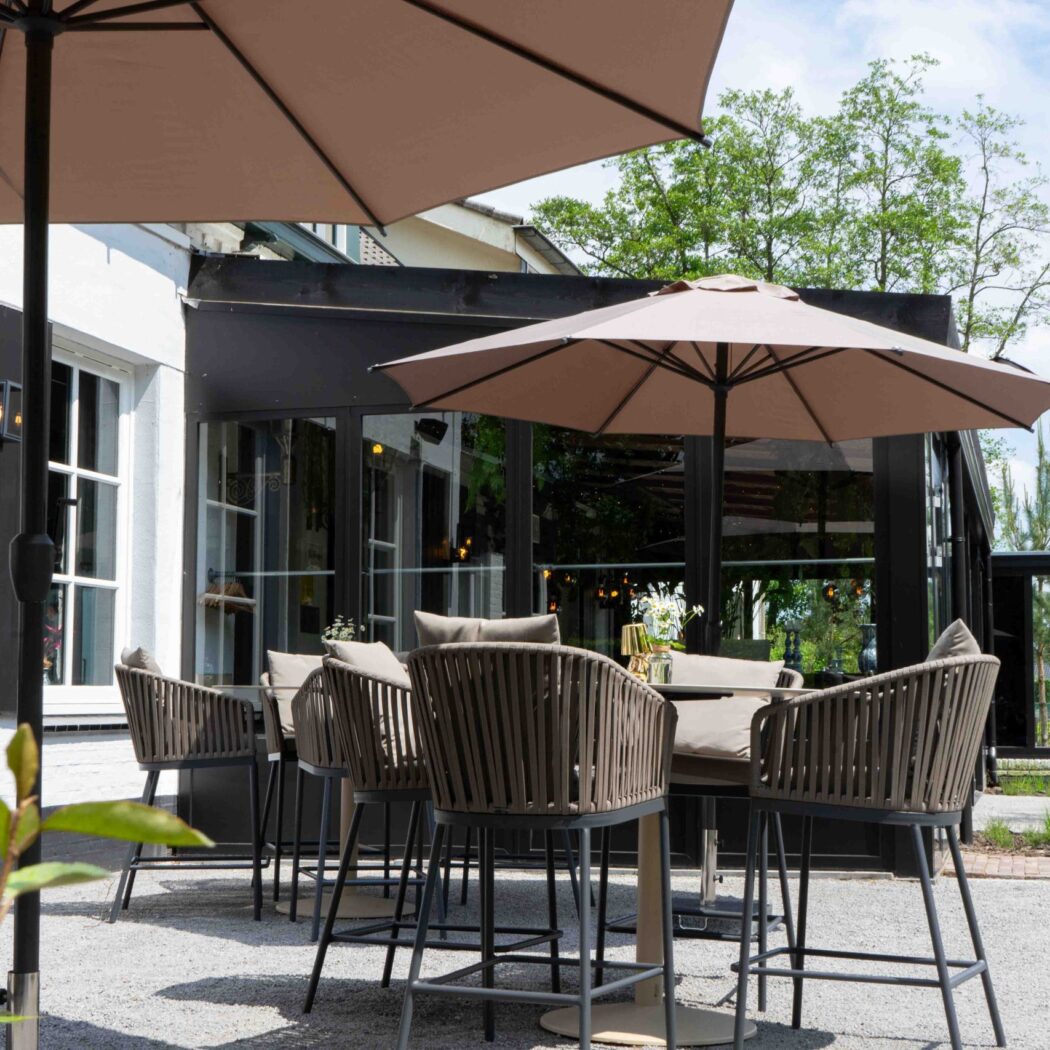 Terras met hoge, gevlochten stoelen, tafels en beige parasols buiten een restaurant.