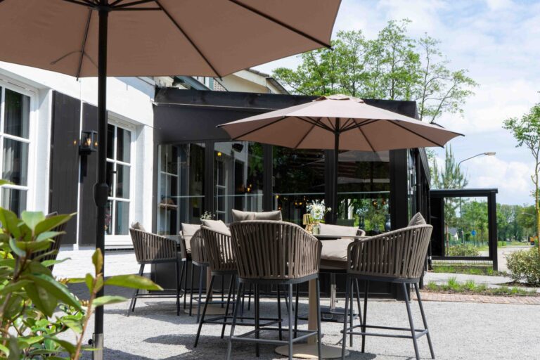 Terras met hoge, gevlochten stoelen, tafels en beige parasols buiten een restaurant.