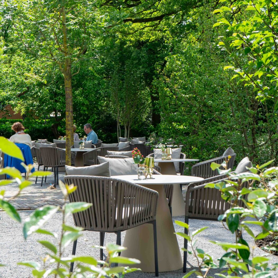 Modern buitenterras met gevlochten stoelen en tafels in een groene tuinomgeving, met gasten.