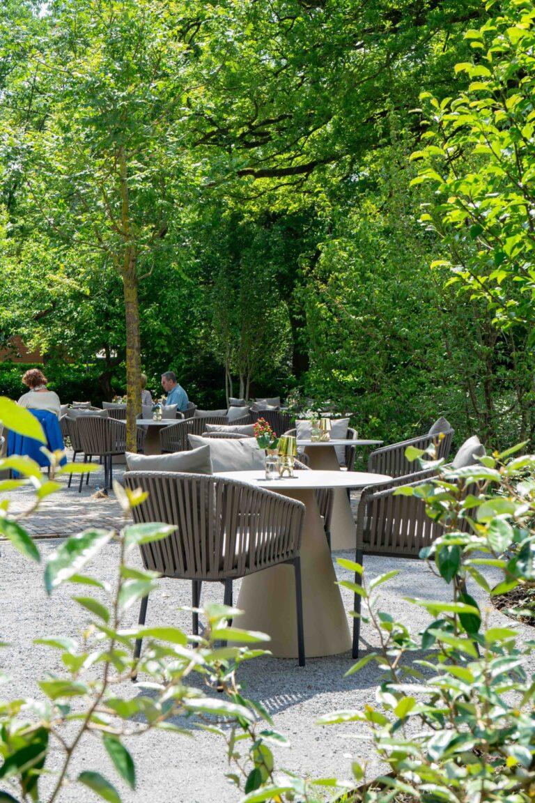 Modern buitenterras met gevlochten stoelen en tafels in een groene tuinomgeving, met gasten.