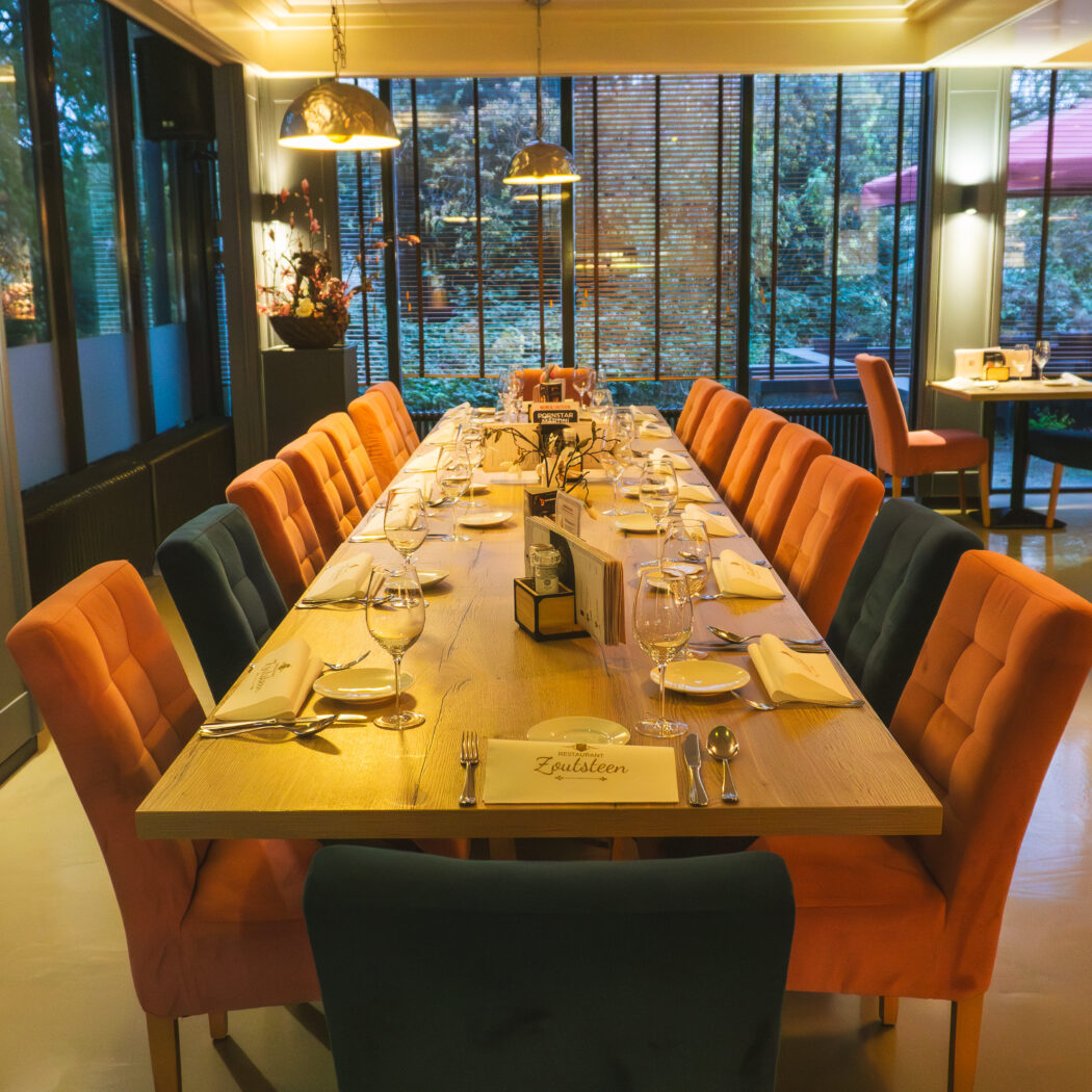 Lange houten eettafel in restaurant, gedekt voor gasten met oranje en donkerblauwe stoelen. Plaatskaarten tonen Zoutsteen.