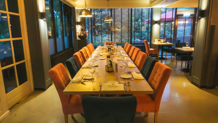 Lange houten eettafel in restaurant, gedekt voor gasten met oranje en donkerblauwe stoelen. Plaatskaarten tonen Zoutsteen.