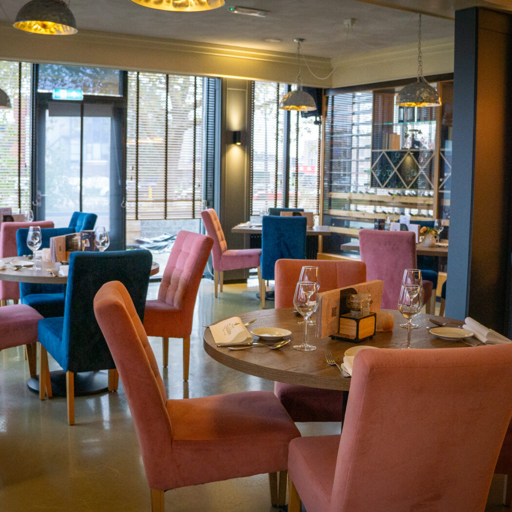 Sfeervol restaurantinterieur met gedekte ronde houten tafels, roze en blauwe stoelen en grote ramen.