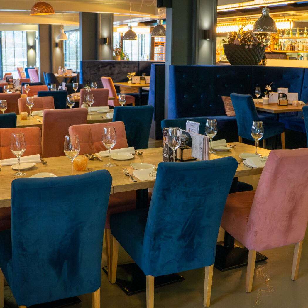Warm ingericht restaurant met gedekte lichte houten tafels, donkerblauwe banken en roze en petrolkleurige stoelen.