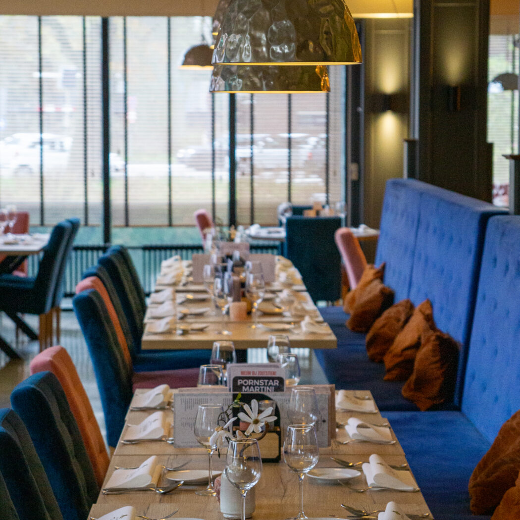 Lange, gedekte restauranttafels met blauwe en oranje fluwelen stoelen, blauwe banken, gehamerde hanglampen en een Pornstar Martini kaart.