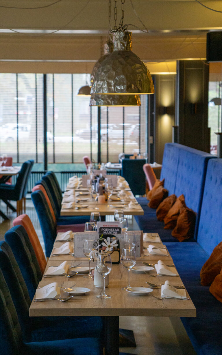 Lange, gedekte restauranttafels met blauwe en oranje fluwelen stoelen, blauwe banken, gehamerde hanglampen en een Pornstar Martini kaart.